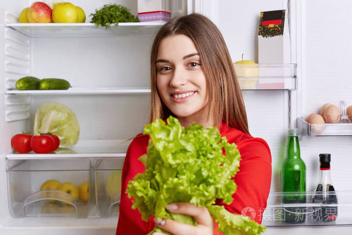 积极的年轻有魅力的女人抱着生菜，素食主义者站在打开的冰箱旁边，装满水果和蔬菜，支持健康的饮食。 可爱的女性站在冰箱附近的厨房里