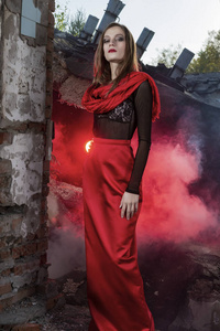 美丽的时尚模特女孩穿着一条红色长裙和一条红色披肩，站在倒塌的建筑中，废墟中升起了红色的烟雾。 概念时尚现代和广告设计。