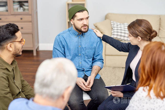 友好的年轻心理学家,手里拿着剪贴板,拍着胡子病人的肩膀,试图鼓励他