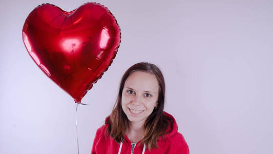 一个穿红色运动衫的女孩手里拿着一个红色的气球，它是一颗心的形式。 学生在白色背景下摆姿势