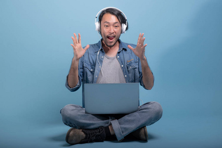年轻的胡须男用蓝色背景的电脑坐在地板上。 惊喜的亚洲年轻潮人使用笔记本电脑听音乐全身射击。 年轻一代嬉皮士工作理念。