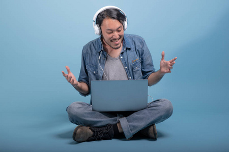 年轻的胡须男用蓝色背景的电脑坐在地板上。 心烦意乱的亚洲年轻人使用笔记本电脑听音乐全身射击。 年轻一代嬉皮士工作理念。
