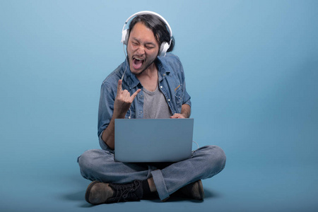 年轻的胡须男用蓝色背景的电脑坐在地板上。 非常兴奋的亚洲年轻人使用笔记本电脑听音乐全身射击。 年轻一代嬉皮士工作理念。