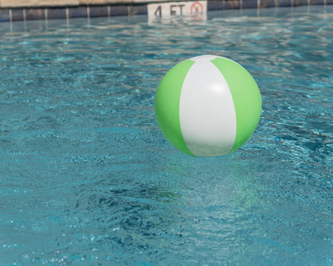 靠近游泳池顶部边缘的亮绿色沙滩球，有深度，没有潜水标志。漂浮在清爽的蓝色海水中，海水在夏季阳光下反射，靠近游泳池顶部甲板和瓷砖
