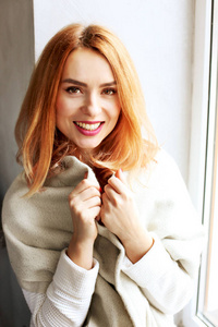 美丽的红头发年轻女子，嘴唇上涂着红色口红，坐在咖啡店的窗户旁，穿着冬秋季节的白色针织毛衣肩膀上的大松围巾。背景，特写..