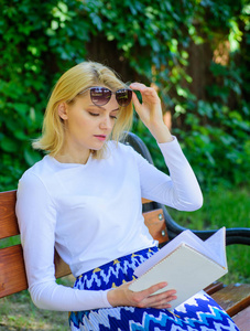 女孩坐板凳放松与书, 绿色自然背景。女金发女郎在公园看书时休息放松。喜欢看书的女孩继续看书。阅读文学作为爱好。书是她的激情