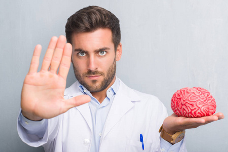 英俊的年轻医生心理学家在灰色的格朗格墙上举着大脑，张开手，用严肃而自信的表情做停止标志，防御手势