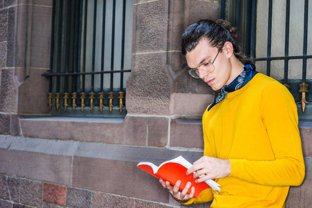 年轻的西班牙裔美国人在纽约市外读红色书籍，戴着眼镜，戴着黄色长袖T恤，脖子上戴着小黑围巾，靠墙站在街上，窗户