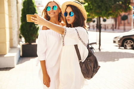两个年轻的女性时尚嬉皮，黑发和金发女性模特在夏天阳光明媚的日子，穿着白色时髦的衣服，在街道背景下的智能手机上为社交媒体拍摄自拍照