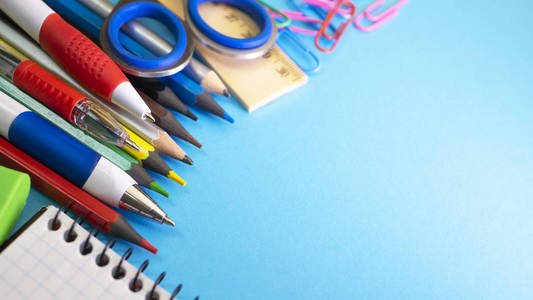 蓝色背景上的学校用品。 钢笔，铅笔，剪刀，尺子，回形针，笔记本和记号笔在桌子上。 上面的视图和复制空间