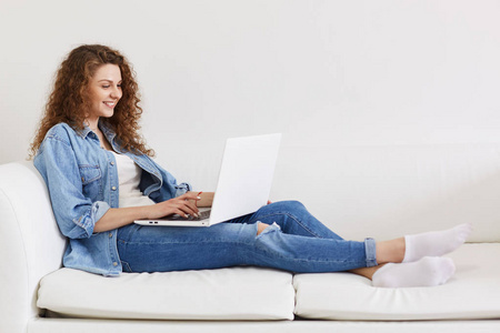 快乐卷曲时尚的女性坐在舒适的白色沙发上享受无线互联网连接，使用笔记本电脑进行在线交流，很高兴收到男朋友的信息。