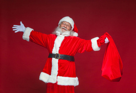 圣诞节。亲切微笑的圣诞老人张开双手向两侧。一方面, 他拿着一个红色的袋子与礼物。在红色背景上被隔离
