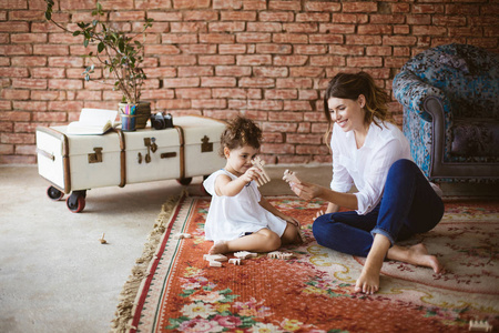 年轻快乐的母亲穿着白色衬衫，小女儿穿着白色连衣裙，在舒适的家中一起在地毯上玩木玩具
