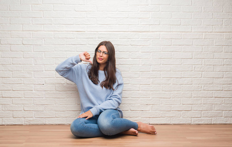 年轻的黑发女人坐在白色砖墙上方的地板上，愤怒的脸上带着消极的迹象，用大拇指表示不喜欢，拒绝的概念