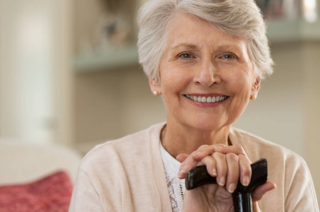 退休妇女带着她的木制手杖在家。 快乐的高级女人在家放松，拿着拐杖，看着相机。 微笑的祖母坐在沙发上。