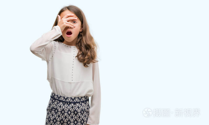 黑发西班牙女孩在震惊中偷看,遮住脸和眼睛,用手透过手指看着尴尬的