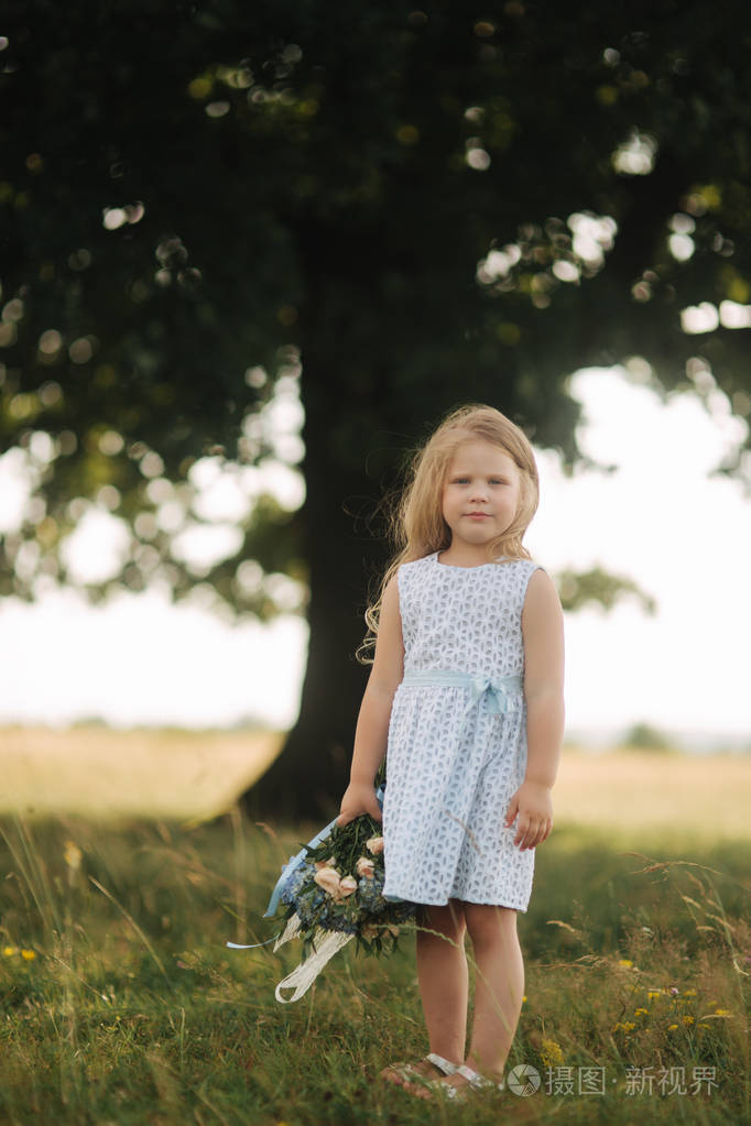 小女孩在天空蓝色礼服与花束站立在领域在大树前面.孩子的微笑和乐趣