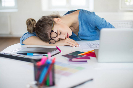 年轻疲惫的女人，头上戴着眼镜，睡在办公桌上，手提电脑，几乎在现代办公室里消磨时间