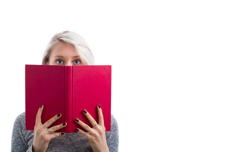 一个漂亮的年轻女孩的肖像隐藏在一本打开的红色书后面，看着害怕的镜头孤立在白色的背景上。