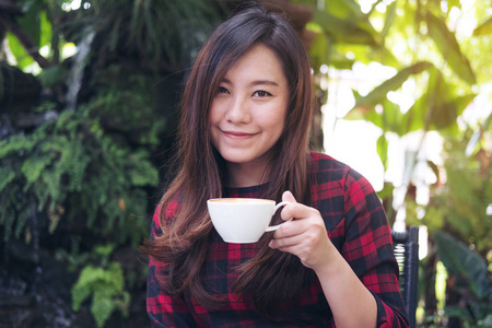 一个美丽的亚洲女人拿着一个白色的杯子喝着热咖啡，在绿色的自然和瀑布花园的背景下感到快乐。