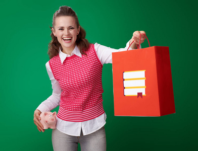 微笑的现代学生妇女穿着红色背心，带着储蓄罐，带着书的购物袋，拿着绿色背景的储蓄罐