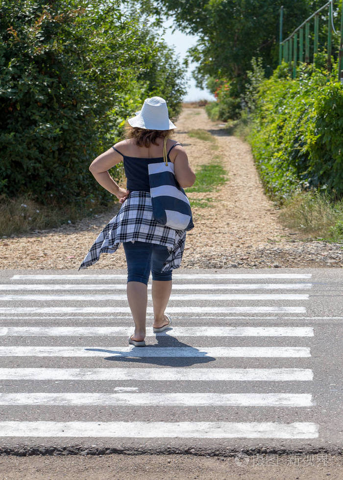 在阳光明媚的天气里戴着帽子和眼镜的丰满女人在行人过路处穿过马路一