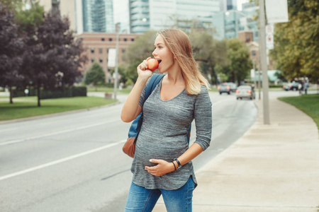 快乐健康的怀孕。 白种人怀孕的年轻金发白种人女人吃苹果的肖像。 美丽的苗条运动期待妈妈女士在繁忙的城市外。