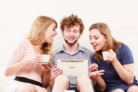 朋友们在平板电脑上浏览上网。年轻人男人和女人坐在沙发上在家放松。