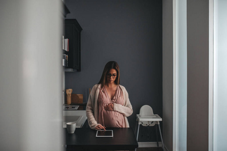 一位孕妇早上在灯光昏暗的厨房里检查她的平板电脑。