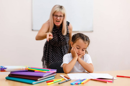 愤怒的老师大喊大叫。 老师对着小女学生尖叫。 小女孩闭着眼睛坐在教室的一张桌子旁