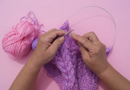 s hands knit a cap of purple yarn. Pink background. Favorite hob