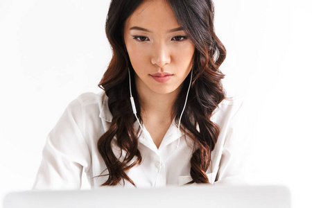 一张严肃的亚洲女人20多岁的照片，穿着办公室服装和耳机坐在桌子上，看着工作室里白色背景上孤立的笔记本电脑