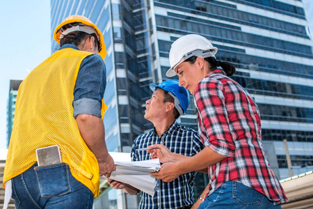 三名工业工程师戴安全帽，工程工作，并与外部建筑的图纸检查交谈。 工程工具和施工理念。 副本空间