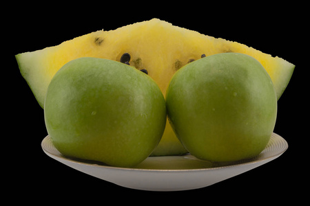 盘子里的黄色西瓜和绿色苹果。 静止的生命被孤立在黑色的背景上。