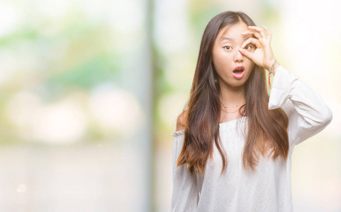 年轻的亚洲女人在孤立的背景下做OK手势，震惊的脸，眼睛透过手指看。 不相信的表情。