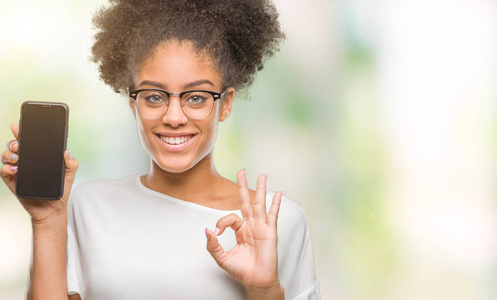 年轻美丽的非洲美国人显示智能手机屏幕在孤立的背景上做OK标志与手指优秀的符号