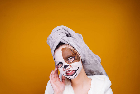 滑稽的年轻女孩在一个白色的特里礼服, 在她的头上的毛巾微笑着, 在她的脸上有一个带有狗脸的保湿面具