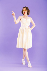 一位身穿夏装站着指着紫罗兰色背景摆姿势的年轻女子的全长画像