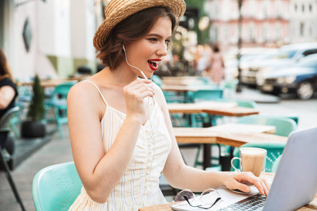 在城市咖啡馆里，坐在桌子旁时，穿着裙子戴着草帽的女人一边用手提电脑视频通话