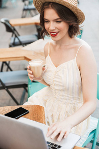 城市咖啡馆里坐在桌子旁，穿着裙子戴着草帽喝着咖啡使用笔记本电脑的微笑女人的垂直形象
