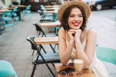 微笑的女人，穿着连衣裙，戴着草帽，一边喝咖啡，一边坐在城市咖啡馆的桌子旁