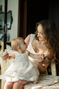 一幅年轻漂亮的母亲在豪迈的室内窗户边和她的女婴玩耍的照片。妈妈穿粉红色衣服