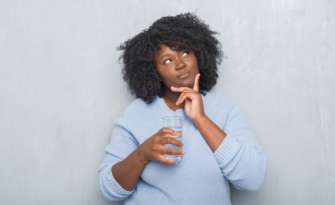 年轻的非洲裔美国妇女在灰色的玻璃墙上喝着一杯水，严肃的脸，思考问题，非常困惑的想法
