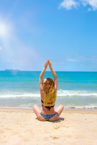 一个年轻漂亮的红头发女孩穿着比基尼在白色沙滩和绿松石海练习瑜伽