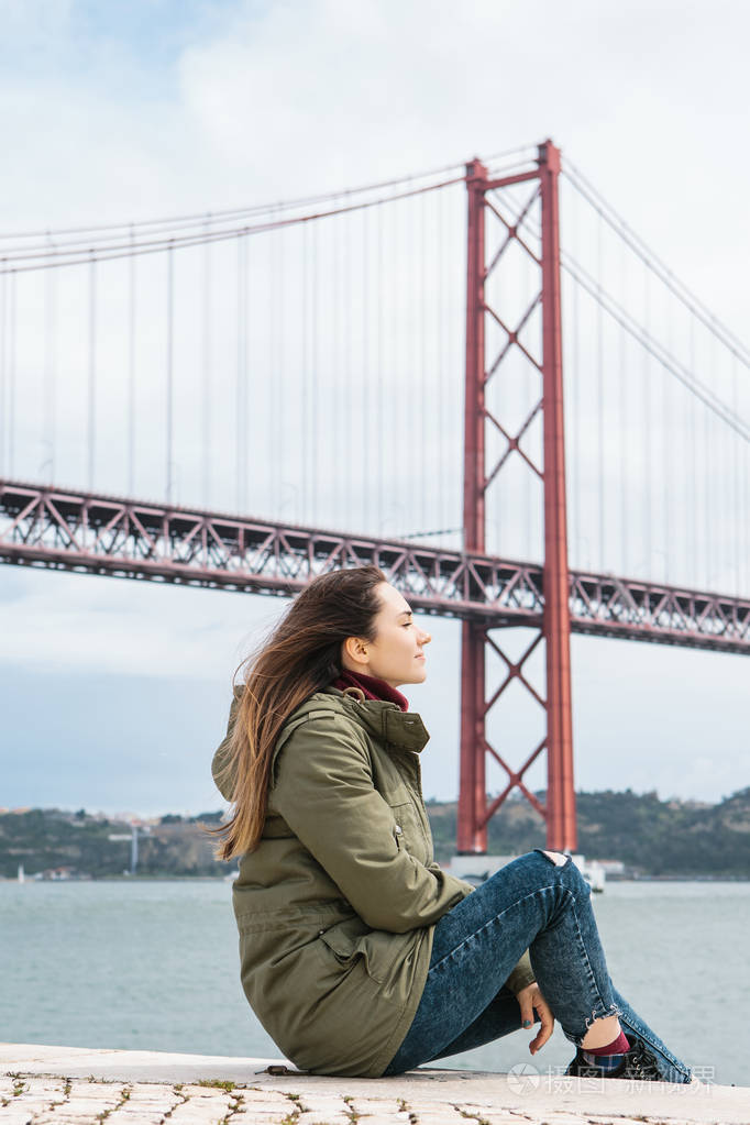 一个孤独的女孩穿着夹克在里斯本的路堤上凉爽的天气坐着凝视着远方和