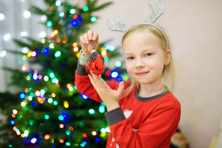 可爱的小女孩用五颜六色的玻璃包装饰圣诞树。 修剪圣诞树。 在家里庆祝圣诞节。