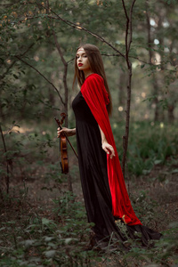迷人的神秘女人，穿着黑色连衣裙和红色斗篷，拉着小提琴在森林里
