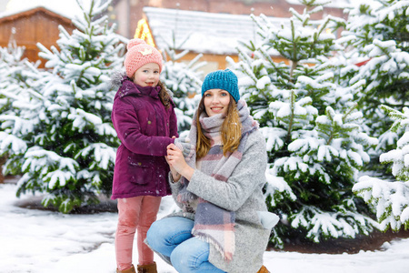 可爱的小女孩和母亲在圣诞树市场上的微笑。愉快的孩子, 女儿和年轻妇女在冬天衣裳选择圣诞节树在圣诞节市场以光在背景上在冬天雪天