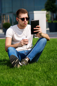 年轻的帅哥戴着太阳镜，使用平板电脑和耳机，喝着咖啡去，坐在草地上，享受现代建筑带来的阳光明媚的一天。男模拍照拍摄，都市时尚生