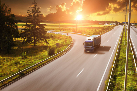 带拖车的蓝色卡车在乡间的道路上行驶，田野和绿树映衬着天空，日落